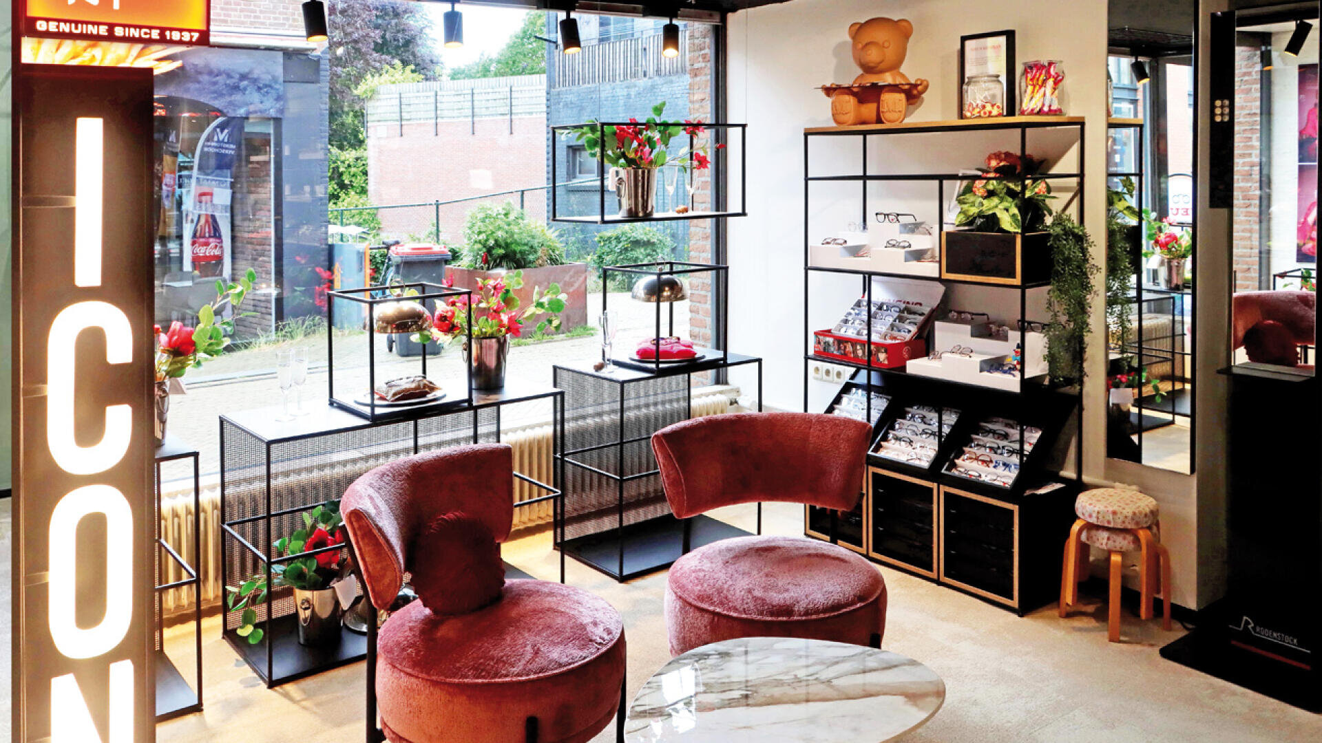 Ein modernes, stilvolles Ladeninterieur mit rosa Samtstühlen, Glasregalen mit Pflanzen und Accessoires, einem Marmortisch und großen Fenstern, die viel Tageslicht hereinlassen. Links leuchtet ein Schild mit der Aufschrift „ICON“.
