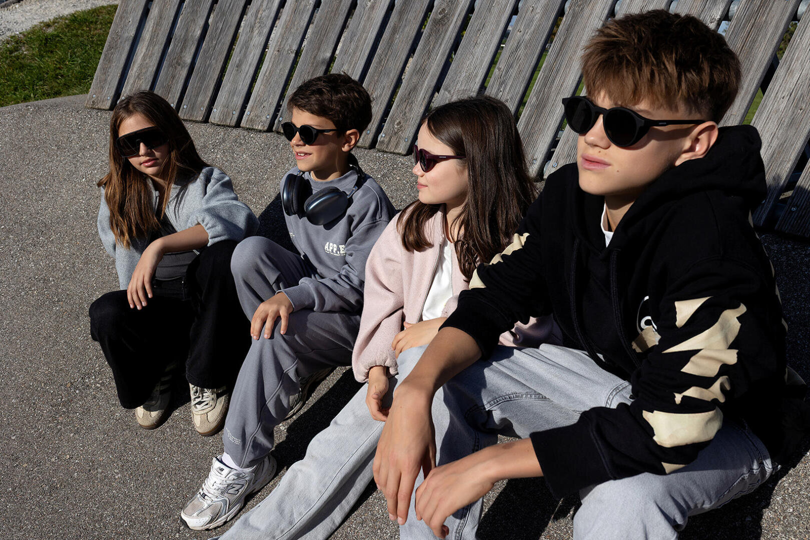 Four teenagers in sunglasses sit on the ground in the sunlight, leaning against a wooden fence. They are dressed casually and look relaxed as they enjoy nature together.