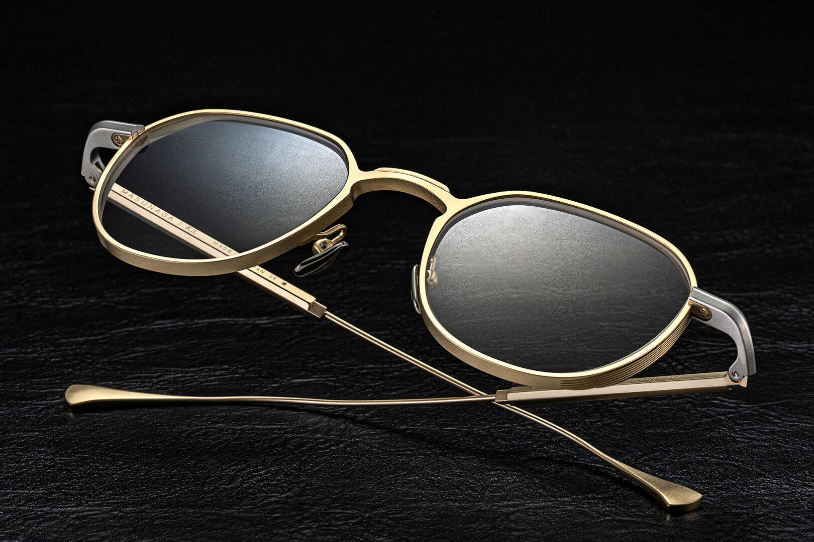 Eine Sonnenbrille mit goldenem Rand und dunklen Gläsern ist auf einer schwarzen, strukturierten Oberfläche abgebildet, wobei die Arme gekreuzt sind und die Gläser nach oben zeigen.