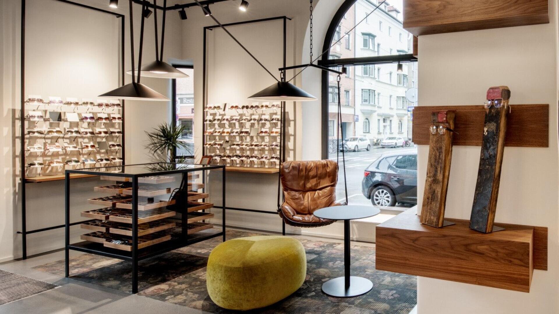 A modern eyewear store interior with display shelves of glasses, a leather chair, a round table, a green ottoman, and large windows showing parked cars and buildings outside. Warm lighting creates a cozy atmosphere.
