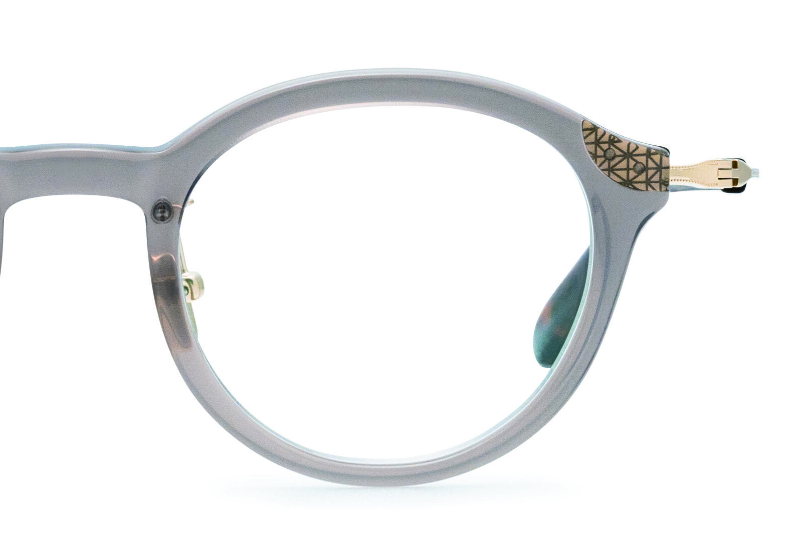 A close-up of a round, light gray eyeglass frame with a gold geometric pattern on the upper right corner and gold hinge accents. The background is white.