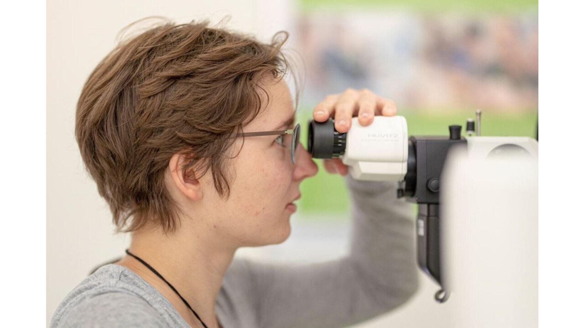 A person with short brown hair and glasses undergoes an eye examination, looking into a medical device used by optometrists in a bright, clinical setting.