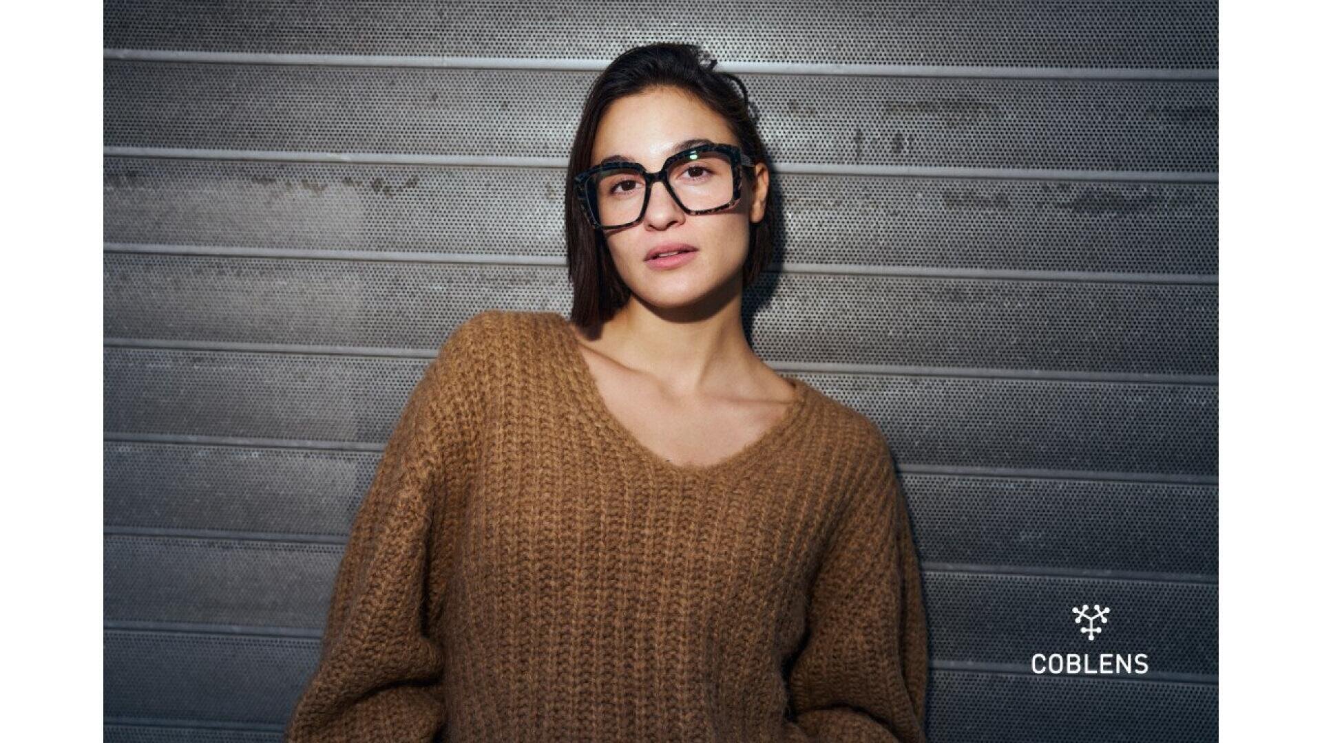 A woman wearing large black glasses and a chunky brown sweater stands against a metallic, textured wall. The COBLENS logo is displayed in the lower right corner.