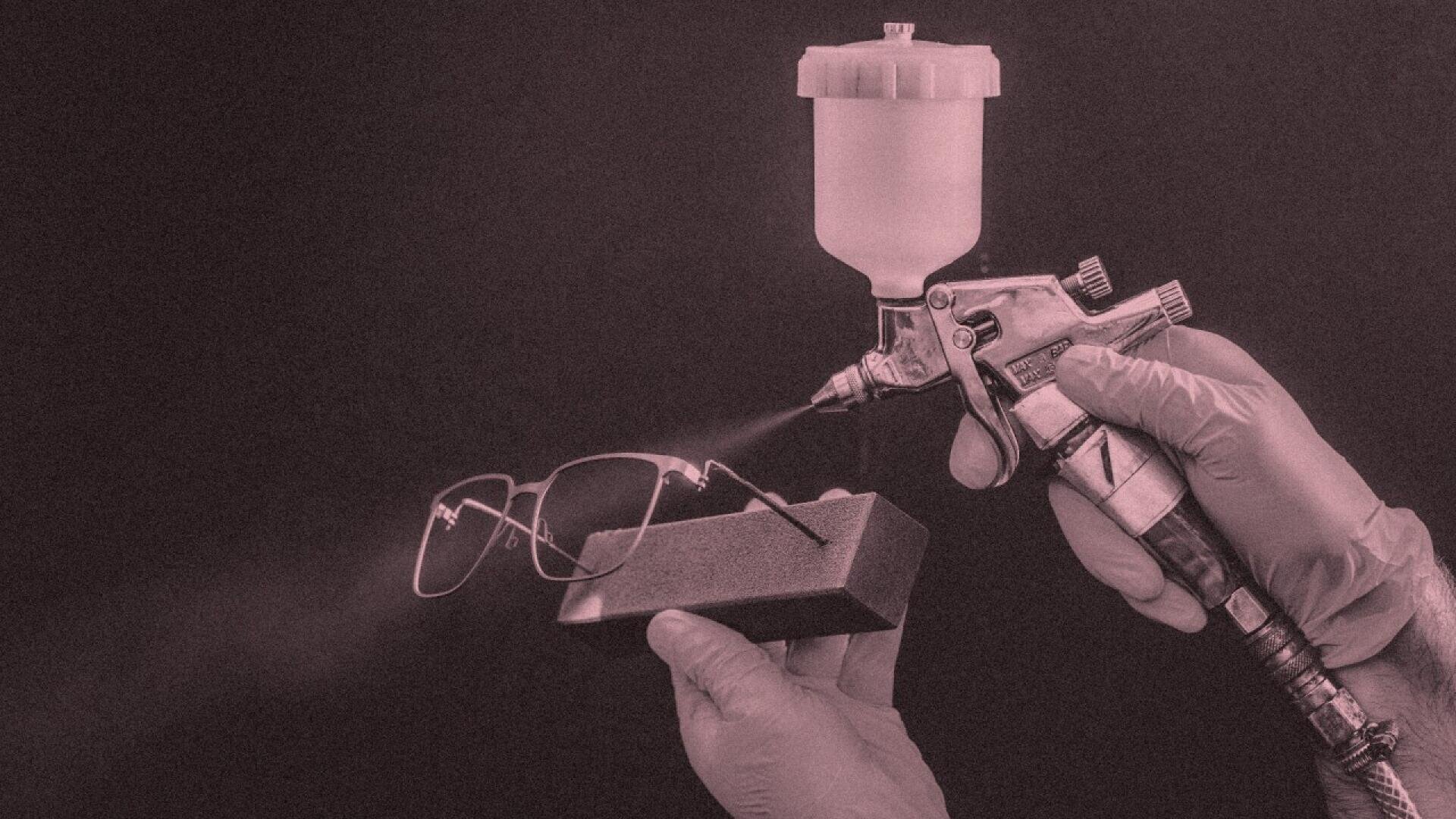 A person wearing gloves uses a spray gun to coat a pair of eyeglasses held in a foam block against a dark background.
