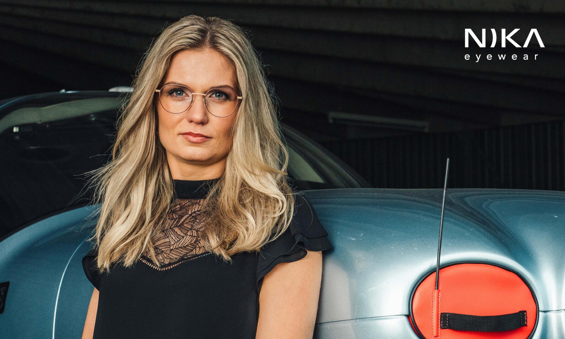 Eine Frau mit langen blonden Haaren und einer runden Brille steht neben einem blauen Auto und trägt ein schwarzes Top mit Spitzenverzierung. Das NIKA-Brillenlogo erscheint in der oberen rechten Ecke.