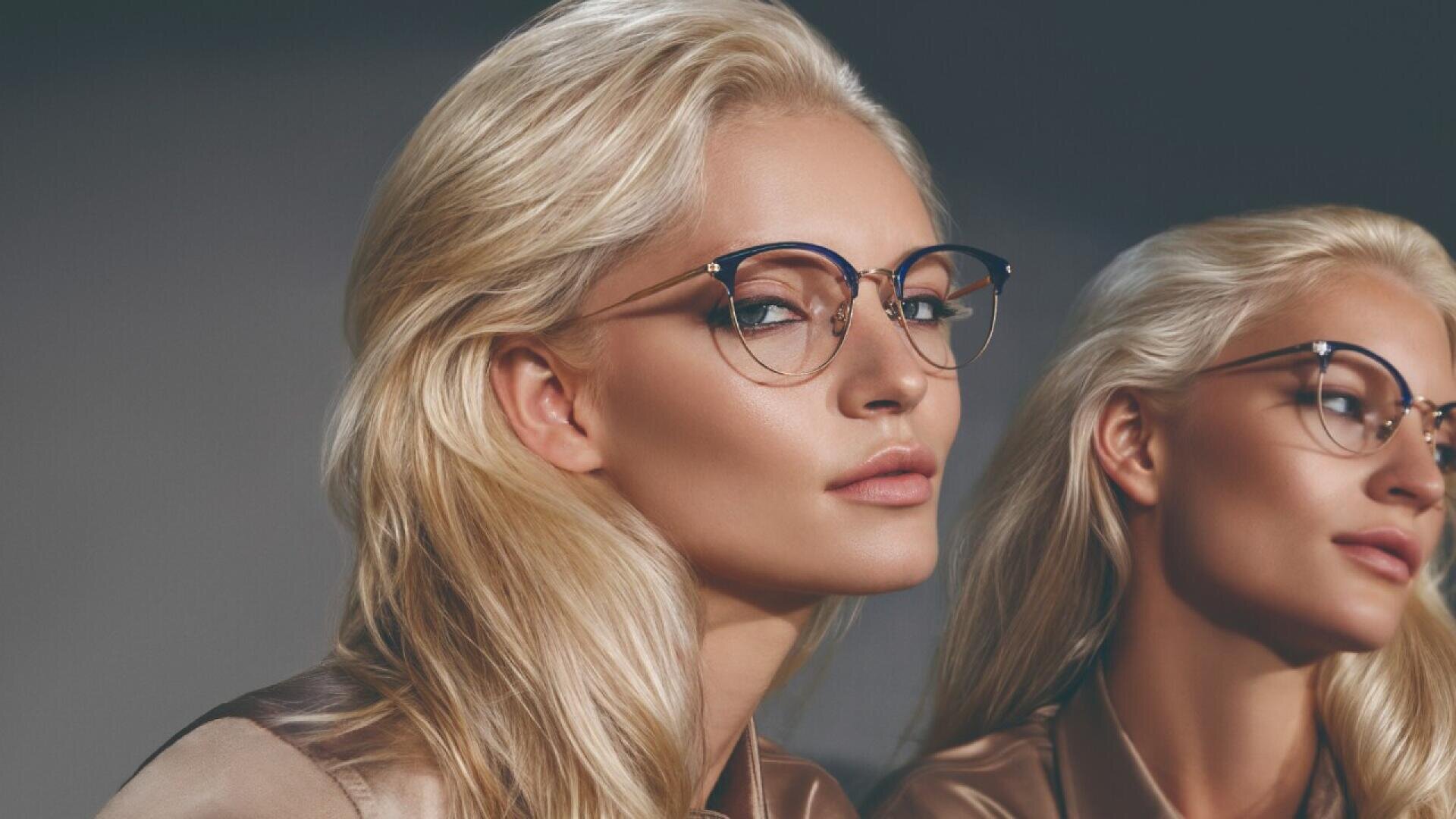 A woman with long blonde hair, wearing round eyeglasses and a silky brown blouse, poses confidently next to her reflection in a mirror against a dark background.