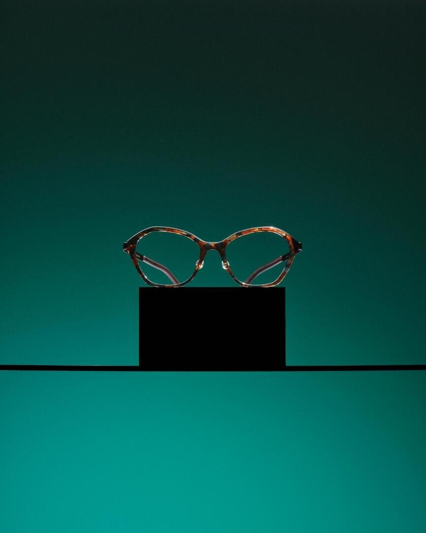 A pair of brown, round eyeglasses is displayed on a black rectangular stand against a gradient teal background, creating a minimalist and elegant composition.