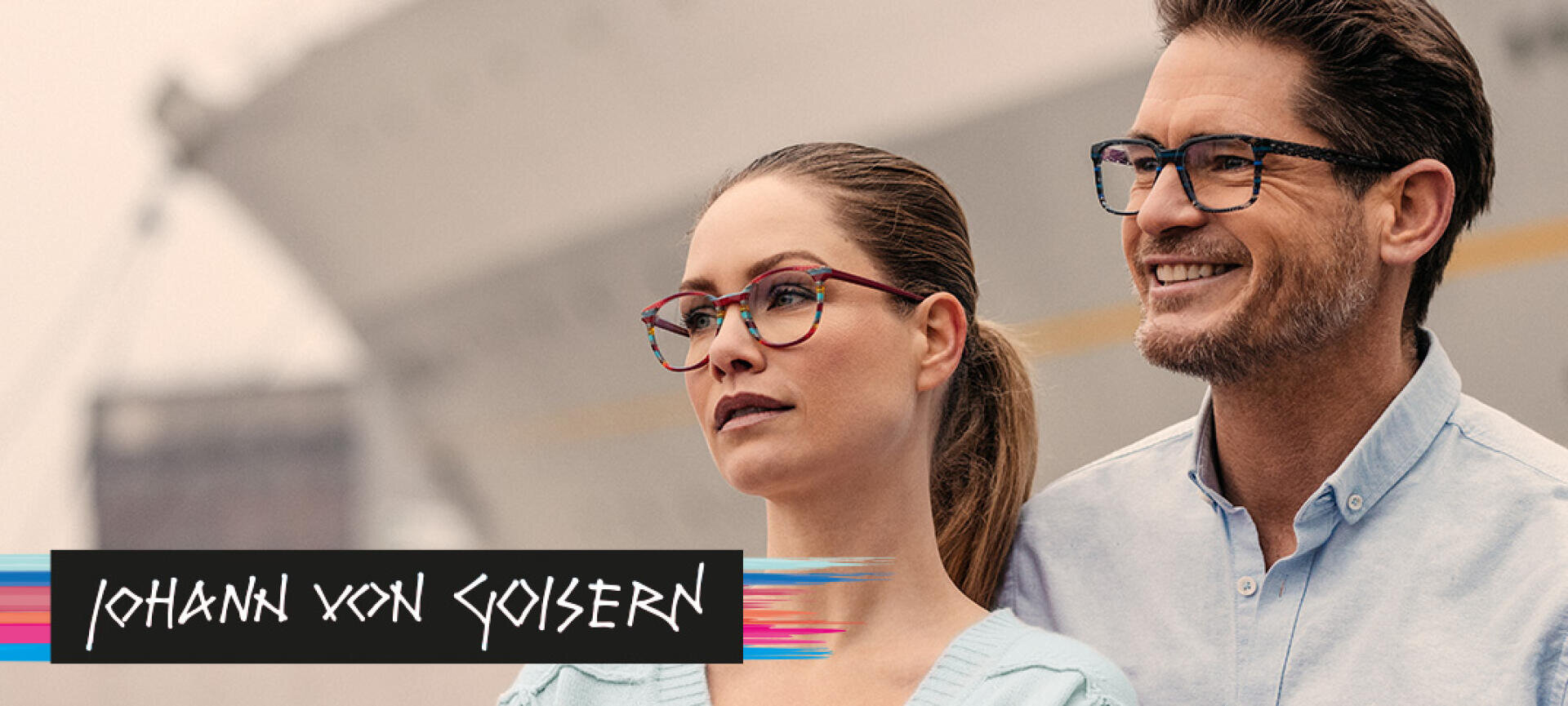 Eine Frau und ein Mann mit modischen Brillen stehen im Freien, beide blicken zur Seite und lächeln. In der unteren linken Ecke des Bildes ist das Logo von Johann von Goisern zu sehen.