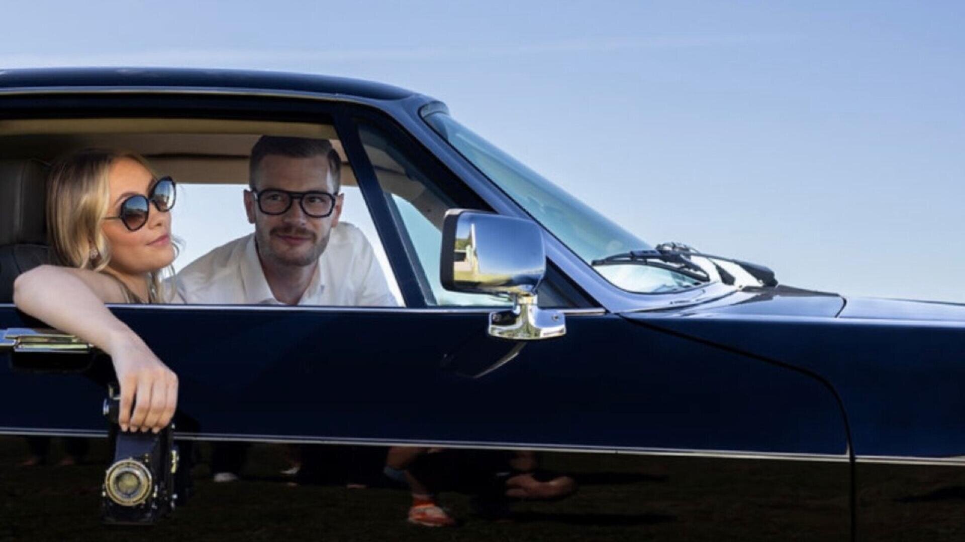 A woman wearing sunglasses and a man with glasses are sitting in the front seats of a classic black car. The woman rests her arm out the window, holding a vintage camera, with a clear blue sky in the background.
