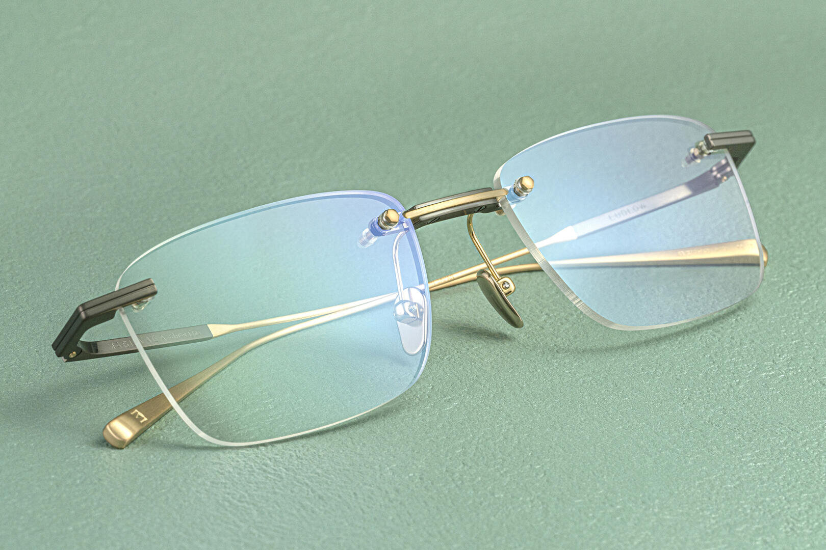 Eine randlose Brille mit goldenen Bügeln und blau getönten Gläsern ruht auf einer strukturierten grünen Oberfläche.