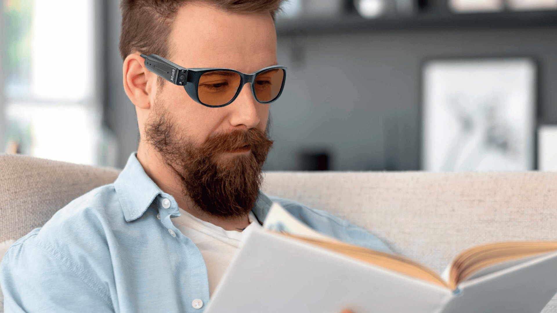 Ein bärtiger Mann mit einer Blaulichtbrille und einem hellblauen Hemd sitzt auf einer Couch und liest ein aufgeschlagenes Buch in einem gut beleuchteten, modernen Wohnzimmer.