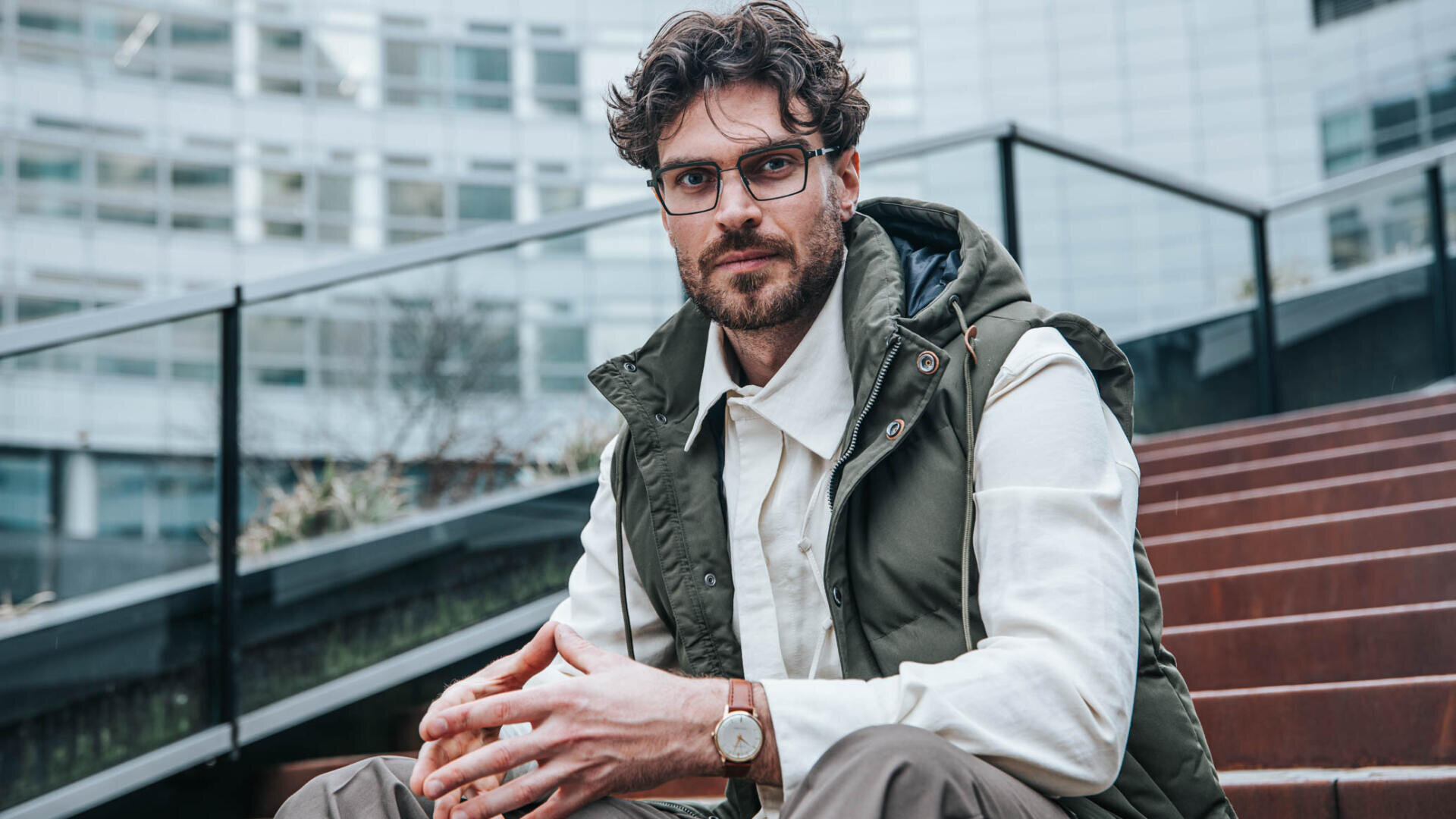 A man with curly hair, a beard and glasses is sitting on a flight of steps. He is wearing a white shirt, an olive green vest, brown trousers and a wristwatch. Modern glass buildings can be seen in the background.