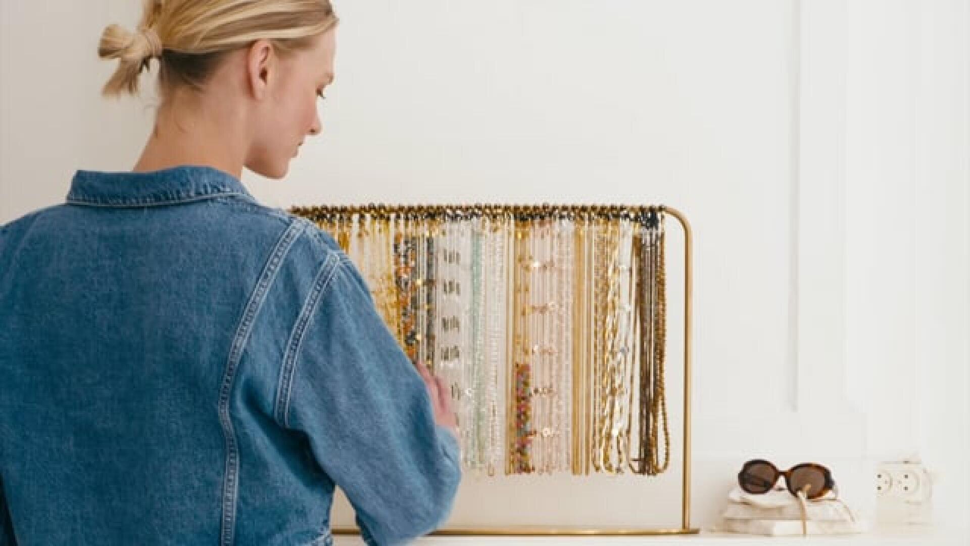 Eine Frau in Jeansjacke steht vor einem Schmuckständer mit bunten Halsketten und Armbändern. Auf der weißen Theke neben ihr liegen Handtücher und eine Sonnenbrille.