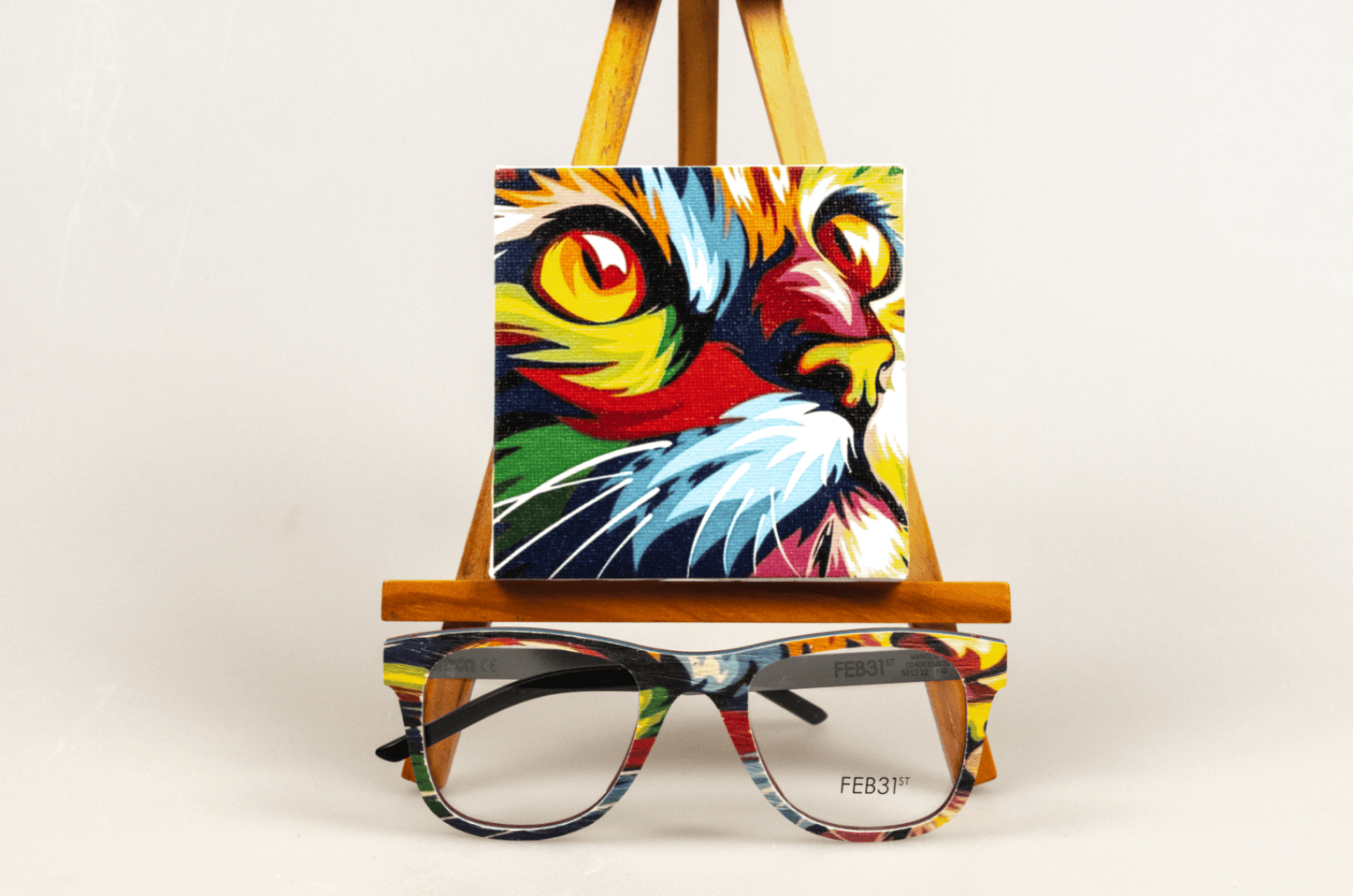 Ein farbenfrohes Gemälde eines Katzengesichts auf einer kleinen Staffelei steht hinter einer Brille mit einem passenden mehrfarbigen Muster. Die Szene hat einen neutralen Hintergrund.