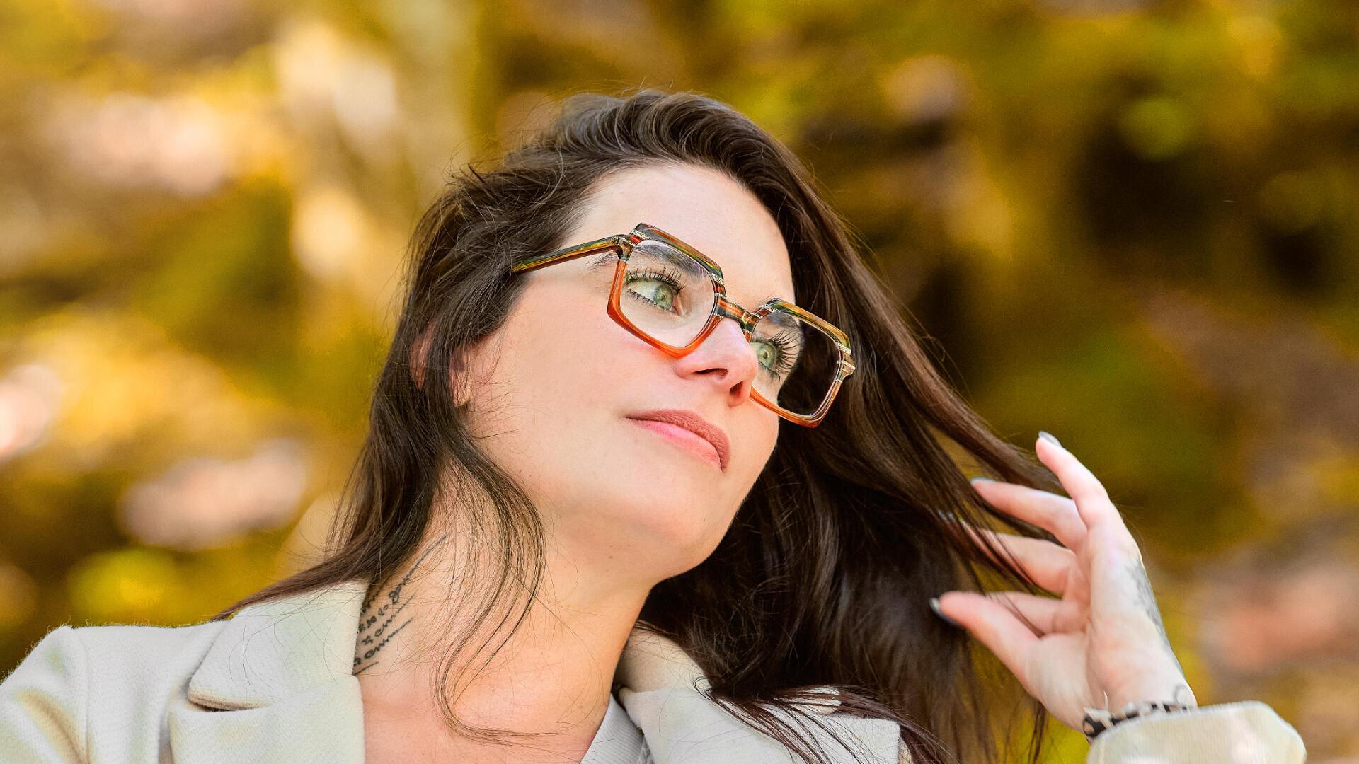Eine Frau mit dunklem Haar und Brille blickt zur Seite und berührt dabei ihr Haar. Sie trägt einen hellen Blazer, der Hintergrund ist in warmen Herbsttönen gehalten.