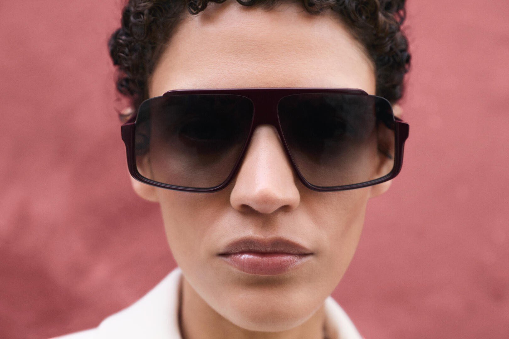 Close-up of a person with short curly hair wearing large, oversized square sunglasses with dark lenses, standing in front of a textured pink wall.