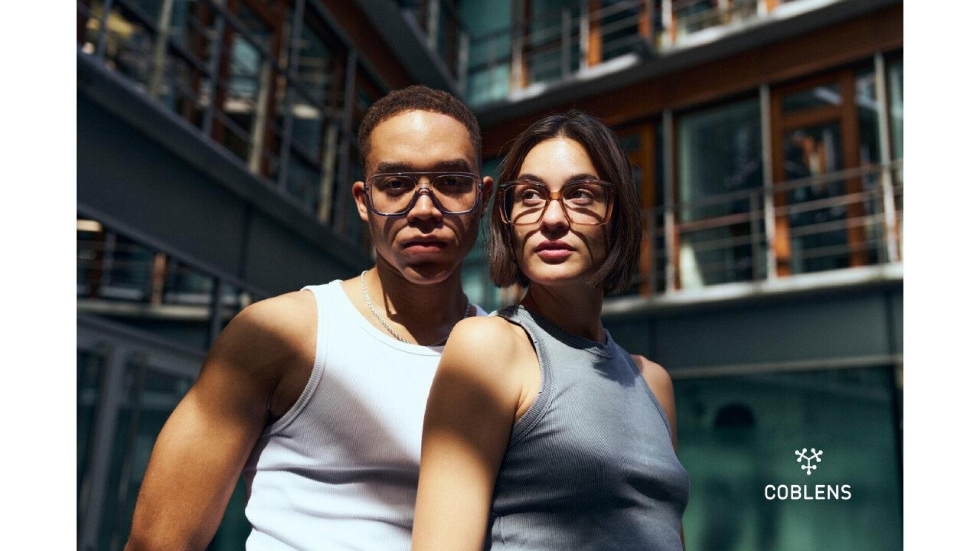 A man and a woman wearing glasses and sleeveless tops stand closely together outdoors, with a modern glass building in the background. The COBLENS logo appears in the lower right corner.