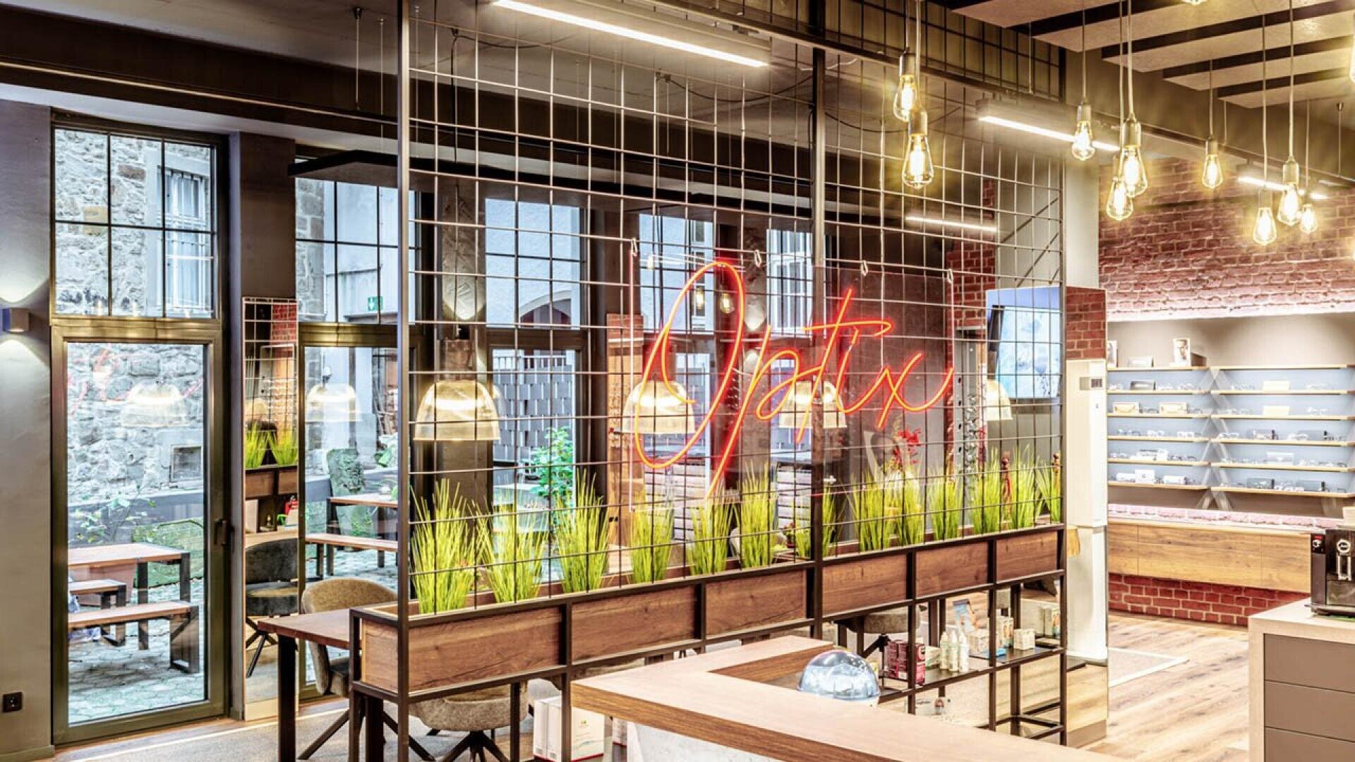 A modern office space with hanging light bulbs, exposed brick walls, large windows, green plants in planters, and a neon sign reading Optix mounted on a wire grid divider.