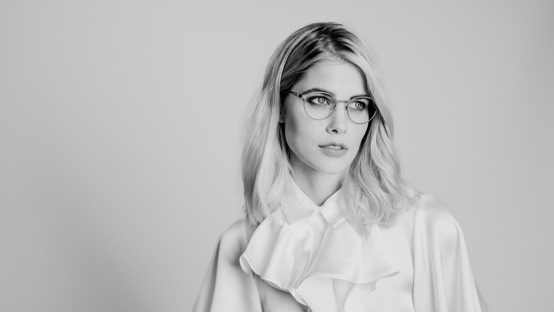 Eine Frau mit hellem Haar, Brille und Rüschenbluse blickt leicht zur Seite. Das Bild ist schwarz-weiß mit schlichtem Hintergrund.
