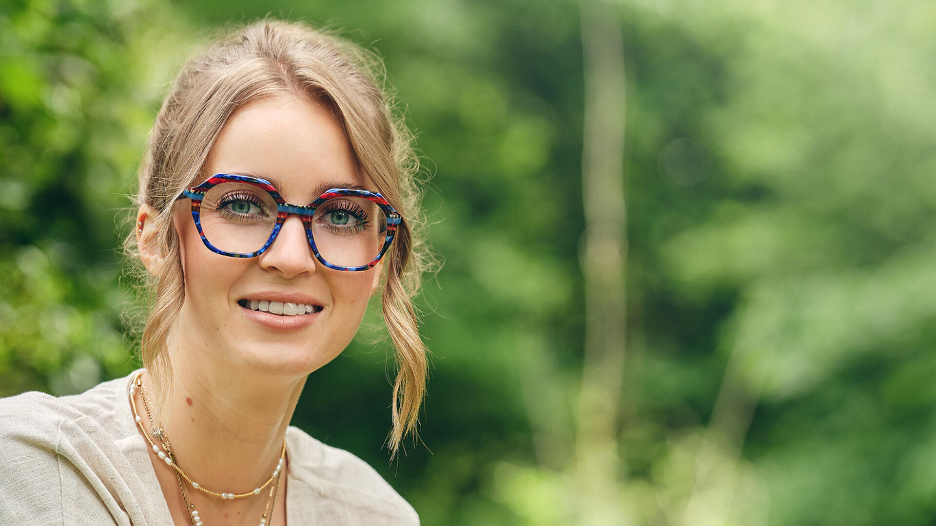 Eine lächelnde Frau mit zurückgebundenem hellem Haar, einer großen, bunten Brille und mehreren Halsketten steht im Freien vor einem verschwommenen grünen Hintergrund.