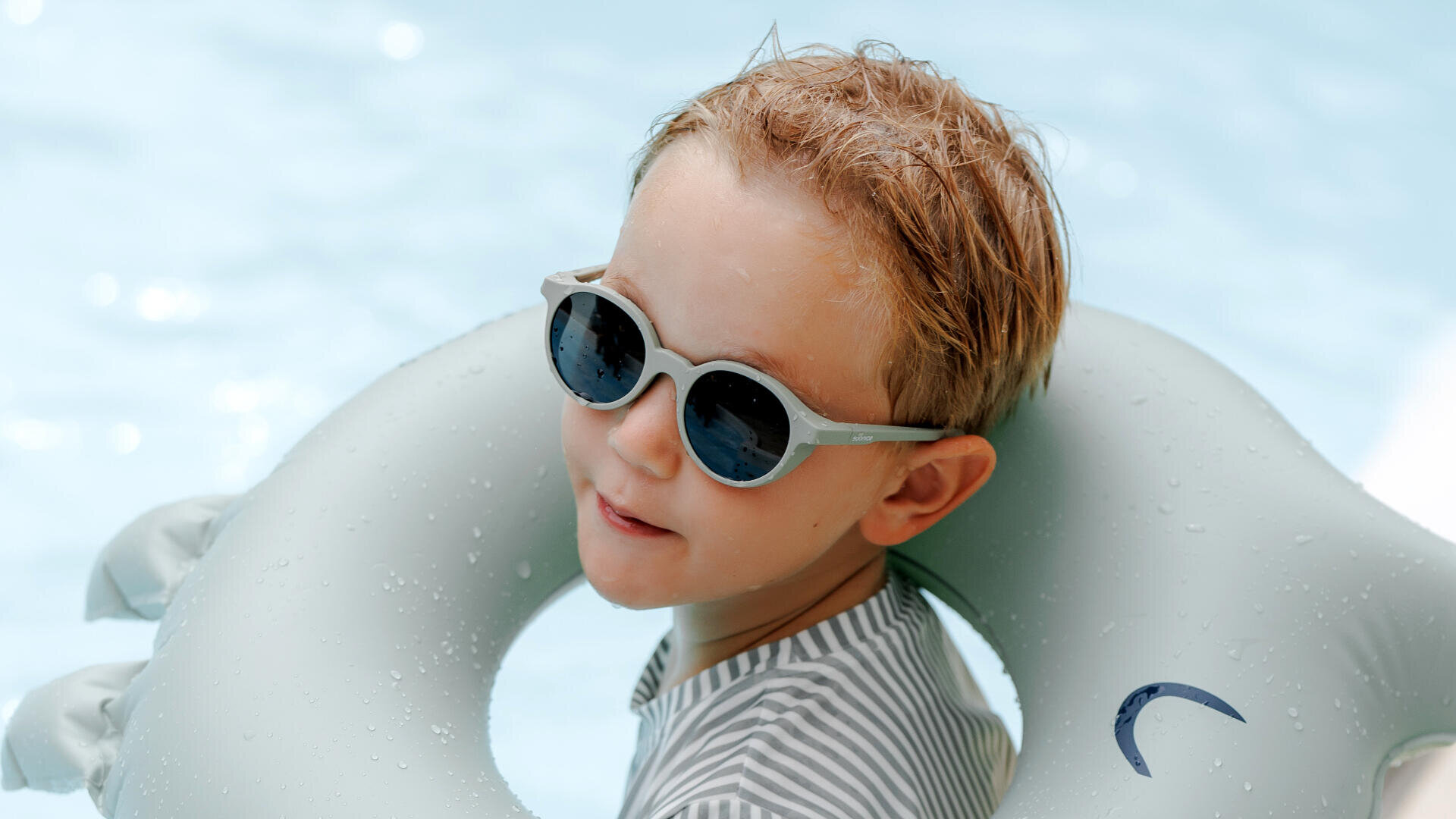 Ein kleiner Junge mit Sonnenbrille und gestreiftem Hemd lächelt in einem Schwimmbecken, umgeben von einem hellgrauen aufblasbaren Schwimmring. Im Hintergrund ist klares blaues Wasser.