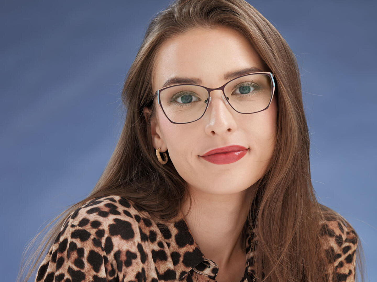 A woman with long brown hair, wearing glasses, gold earrings and a leopard print top, poses in front of a blue background and smiles slightly.