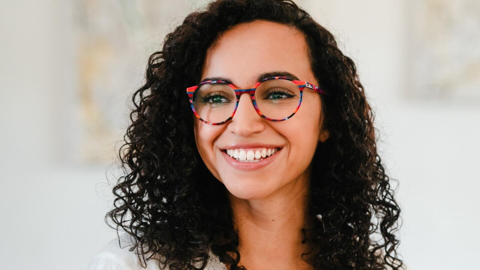 A woman with curly dark hair is smiling and wearing colorful, round eyeglasses. She is indoors with a blurred light background.