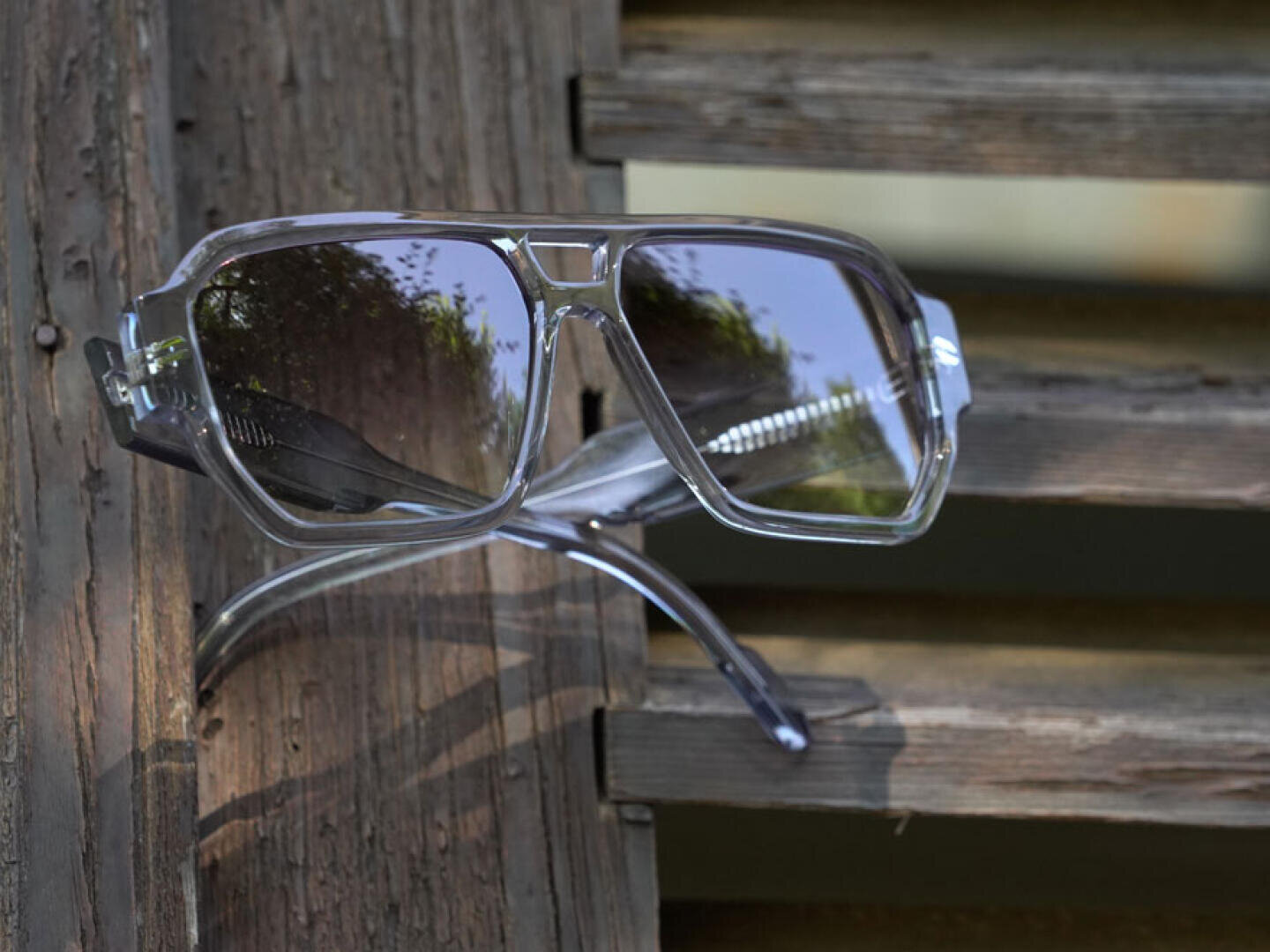 A pair of sunglasses with clear frames hangs from a weathered wooden fence, their lenses reflecting leaves and sunlight.