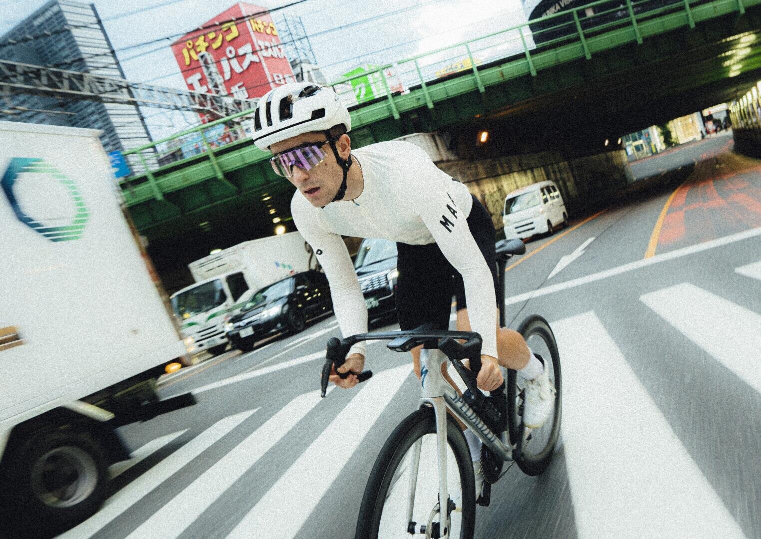 Ein Radfahrer mit weißem Helm, Sonnenbrille und Radfahrerkleidung fährt auf einem Rennrad durch eine Stadtstraße und überholt einen Lastwagen in der Nähe einer Überführung mit japanischen Schildern im Hintergrund.