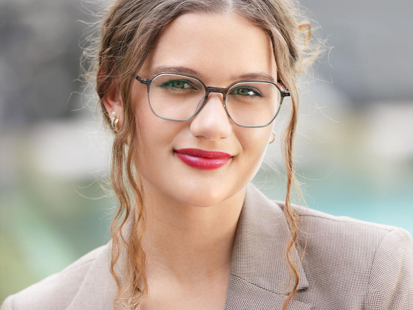 A young woman with wavy light brown hair, glasses and red lipstick wears a beige blazer and smiles slightly at the camera. The background is blurred and outdoors.