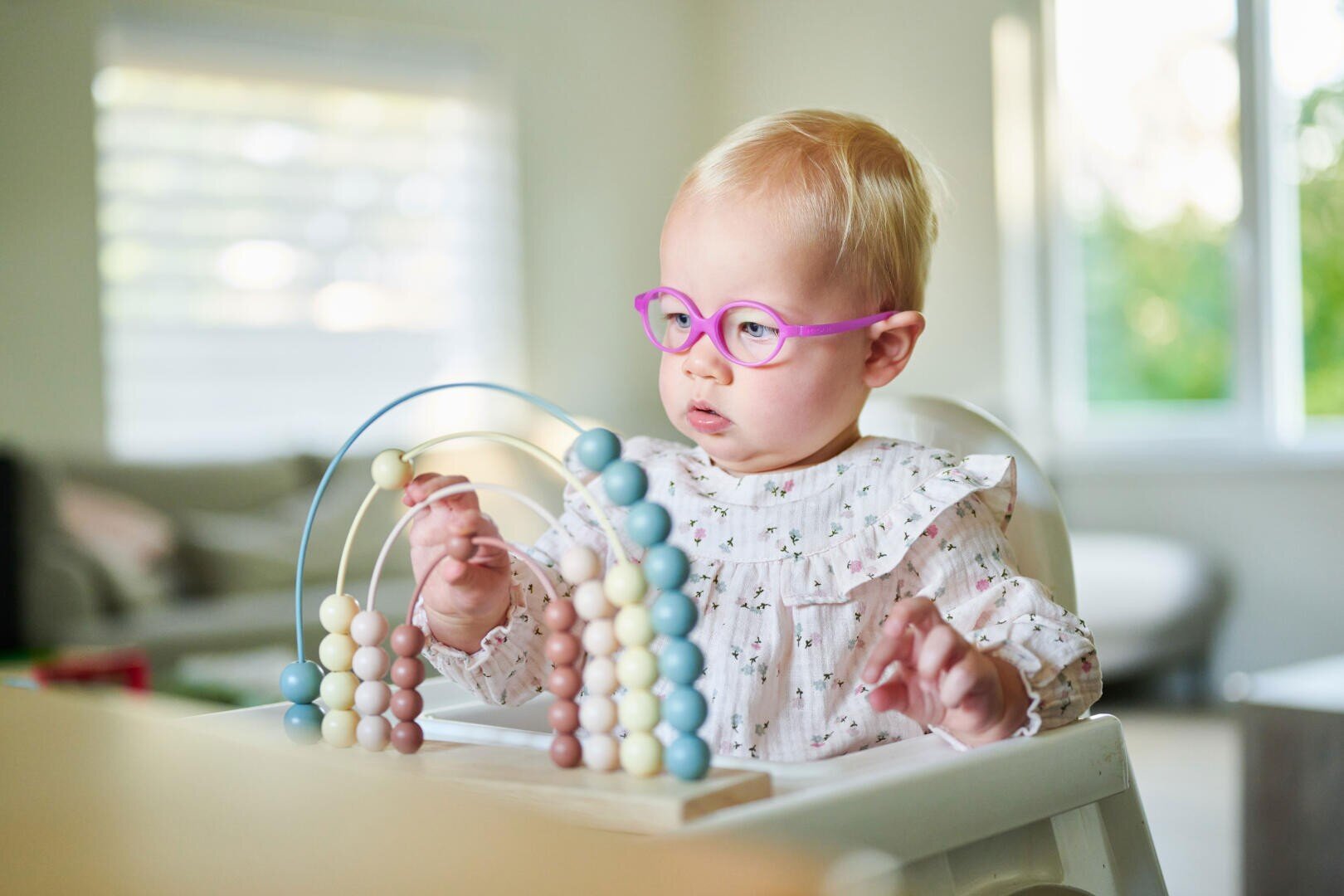 Ein blondes Kleinkind mit rosa Brille sitzt an einem Tisch und spielt mit einem pastellfarbenen Holzabakus. Das Kind ist konzentriert und bewegt die Perlen in einem hellen Raum mit einem Fenster im Hintergrund.