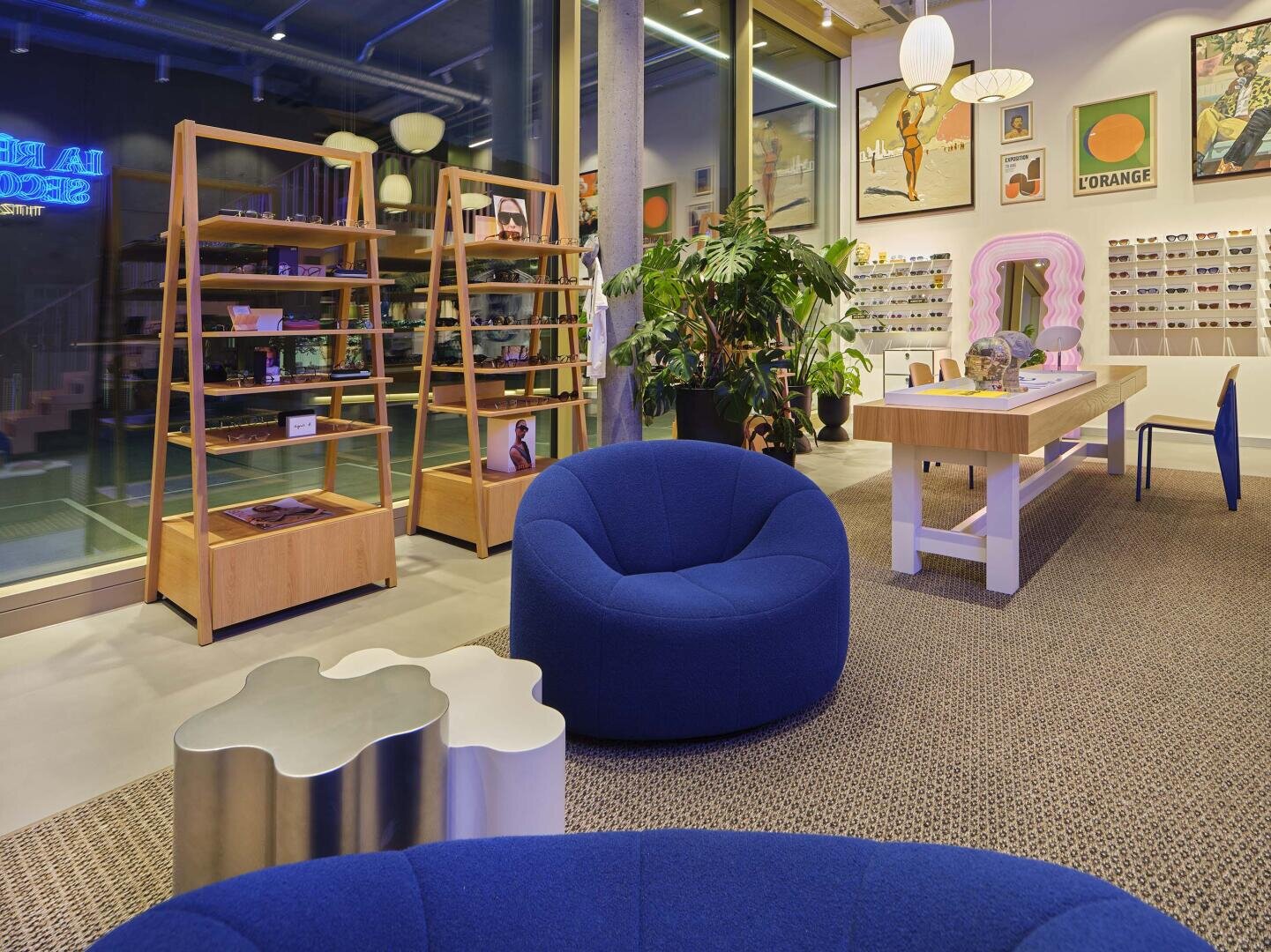 Modernes Brillengeschäft mit blauen Loungesesseln, Holzregalen, Schautischen mit Brillen, Pflanzen, gerahmter Kunst an den Wänden und großen Fenstern, die natürliches Licht hereinlassen.