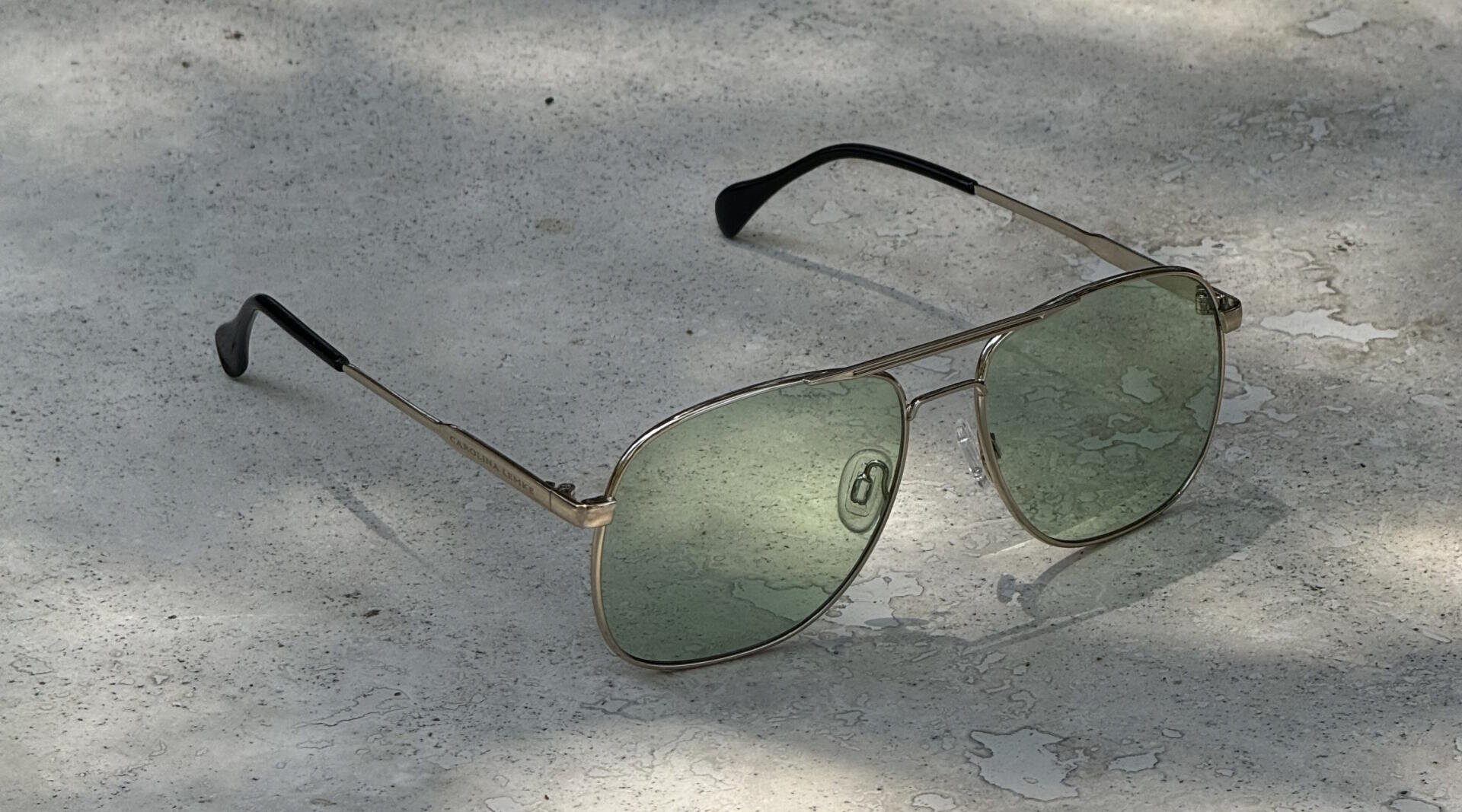 Gold-framed aviator sunglasses with green-tinted lenses rest on a bright, textured concrete surface in partial sunlight and shadow.