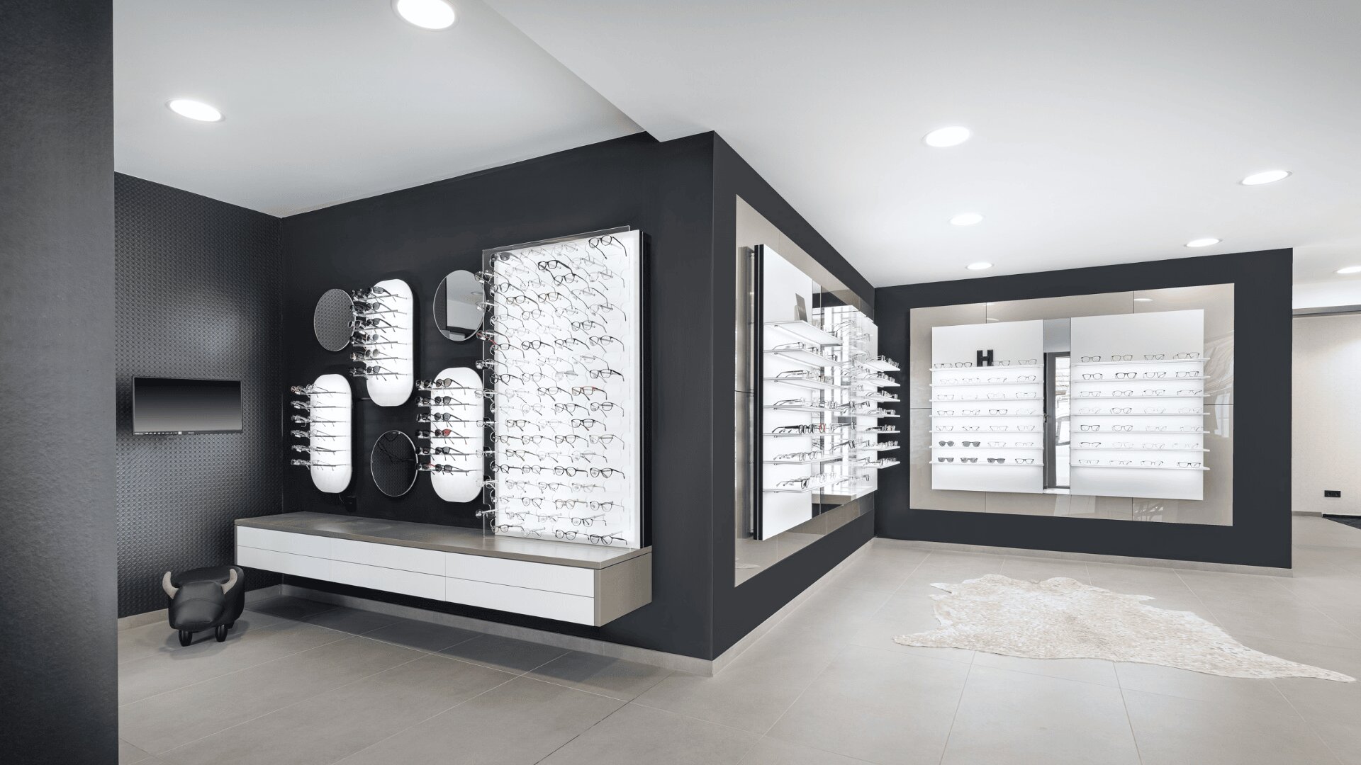 Modernes Brillengeschäft mit schlichtem schwarz-weißem Design, in dem zahlreiche Brillen auf beleuchteten Regalen und Wänden ausgestellt sind. Eine kleine Bank und dekorative Elemente sind sichtbar, die Beleuchtung ist hell und gleichmäßig.
