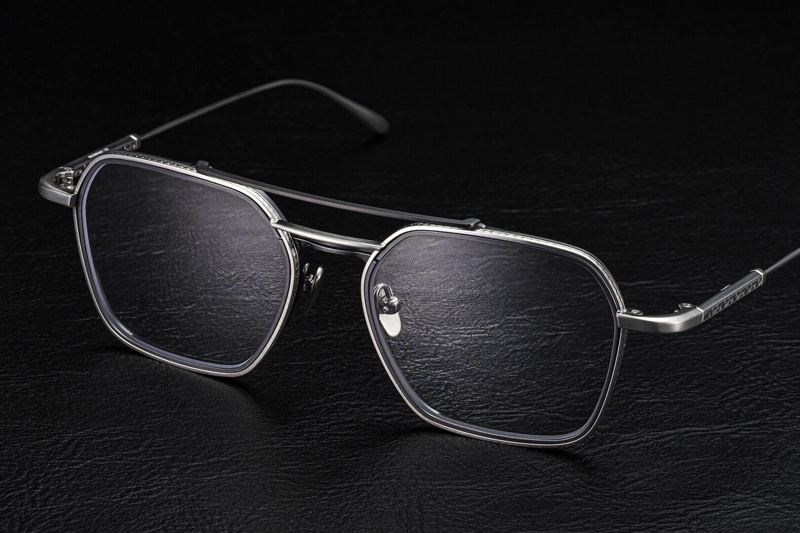 Eine stilvolle, silberne Metallbrille mit großen, rechteckigen Gläsern und dünnen Bügeln, die auf einer schwarzen, strukturierten Oberfläche präsentiert wird.