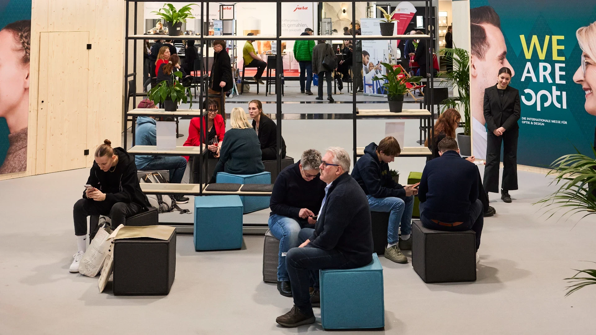 People sit and converse on cube seats in a modern exhibition lounge area, with plants, shelves, and a large WE ARE opti banner in the background. Other attendees walk and browse nearby booths.