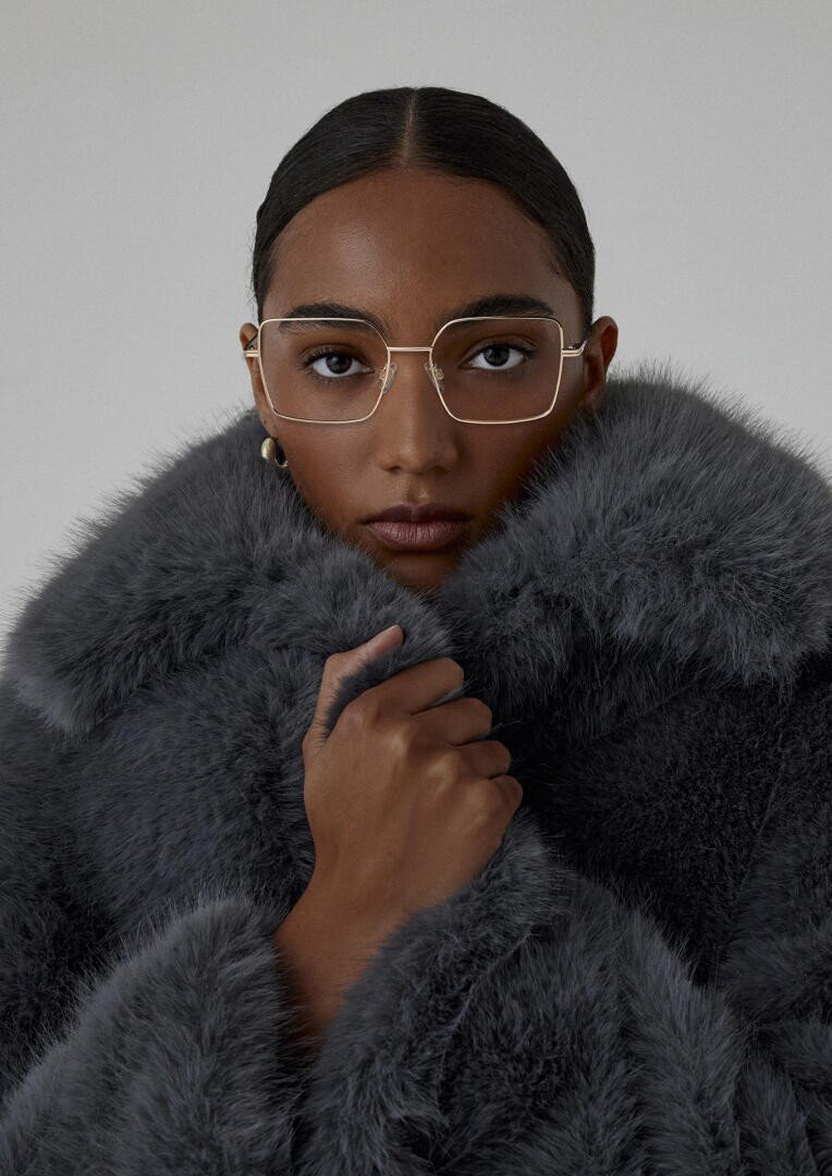 Eine Person mit zurückgegelten Haaren und einer Brille mit goldenen Rändern trägt einen dicken grauen Kunstpelzmantel, hält den Kragen eng am Gesicht und blickt vor einem schlichten Hintergrund direkt in die Kamera.