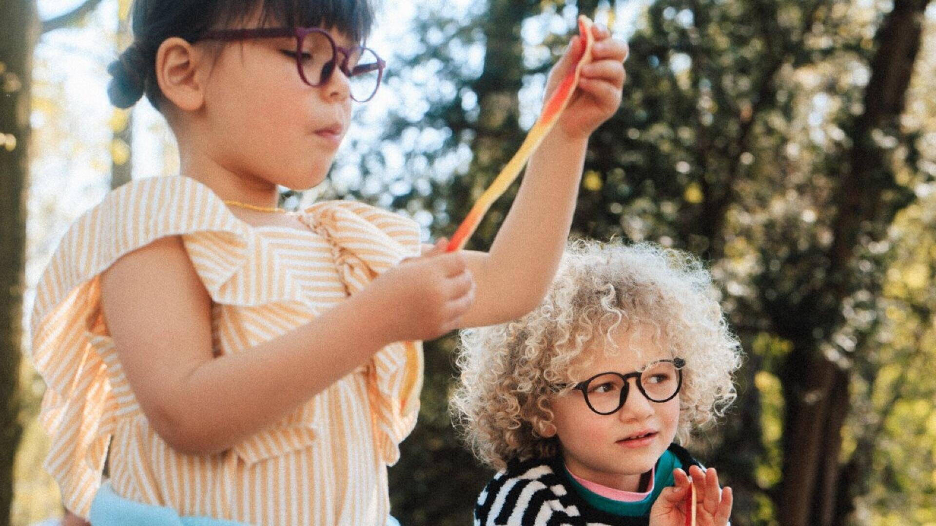 Two young children wearing glasses are outdoors. One is holding a colorful slime toy, while the other watches with interest. Trees and sunlight are visible in the background.