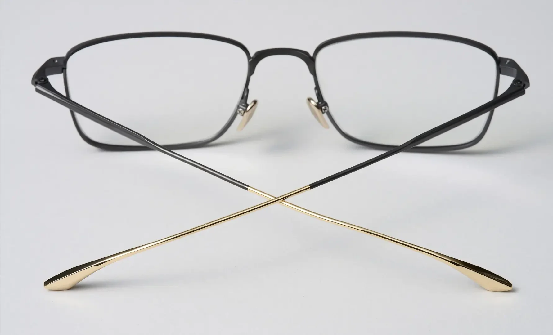 Eine Brille mit schwarzem, rechteckigem Gestell und goldenen Bügeln, dargestellt auf einer weißen Fläche, wobei die Bügel vor dem Gestell in Form eines X gekreuzt sind.