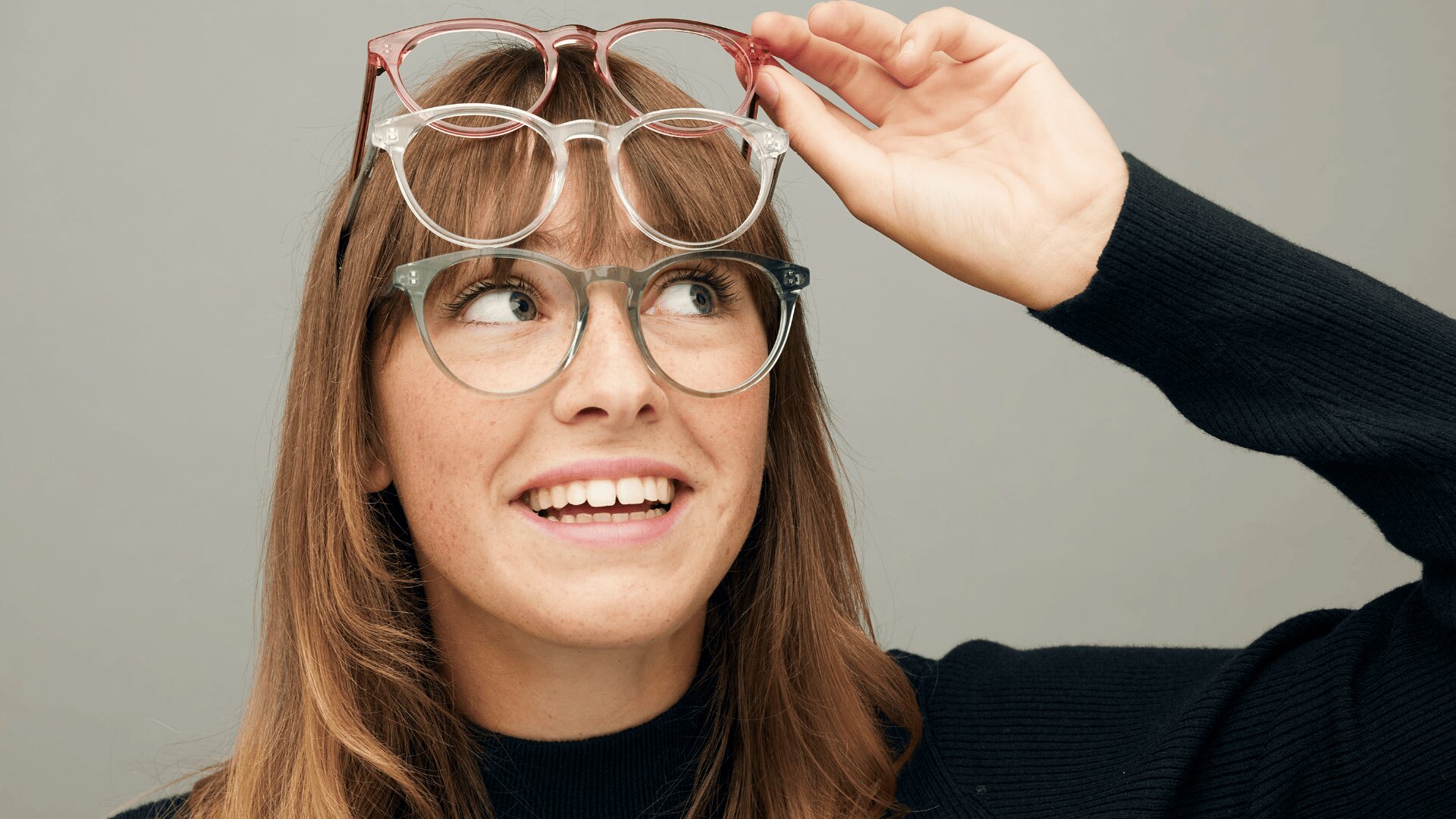 Eine Frau mit langen braunen Haaren und Pony lächelt, während sie zwei Brillen trägt und eine dritte über ihren Kopf hält, vor einem schlichten grauen Hintergrund.