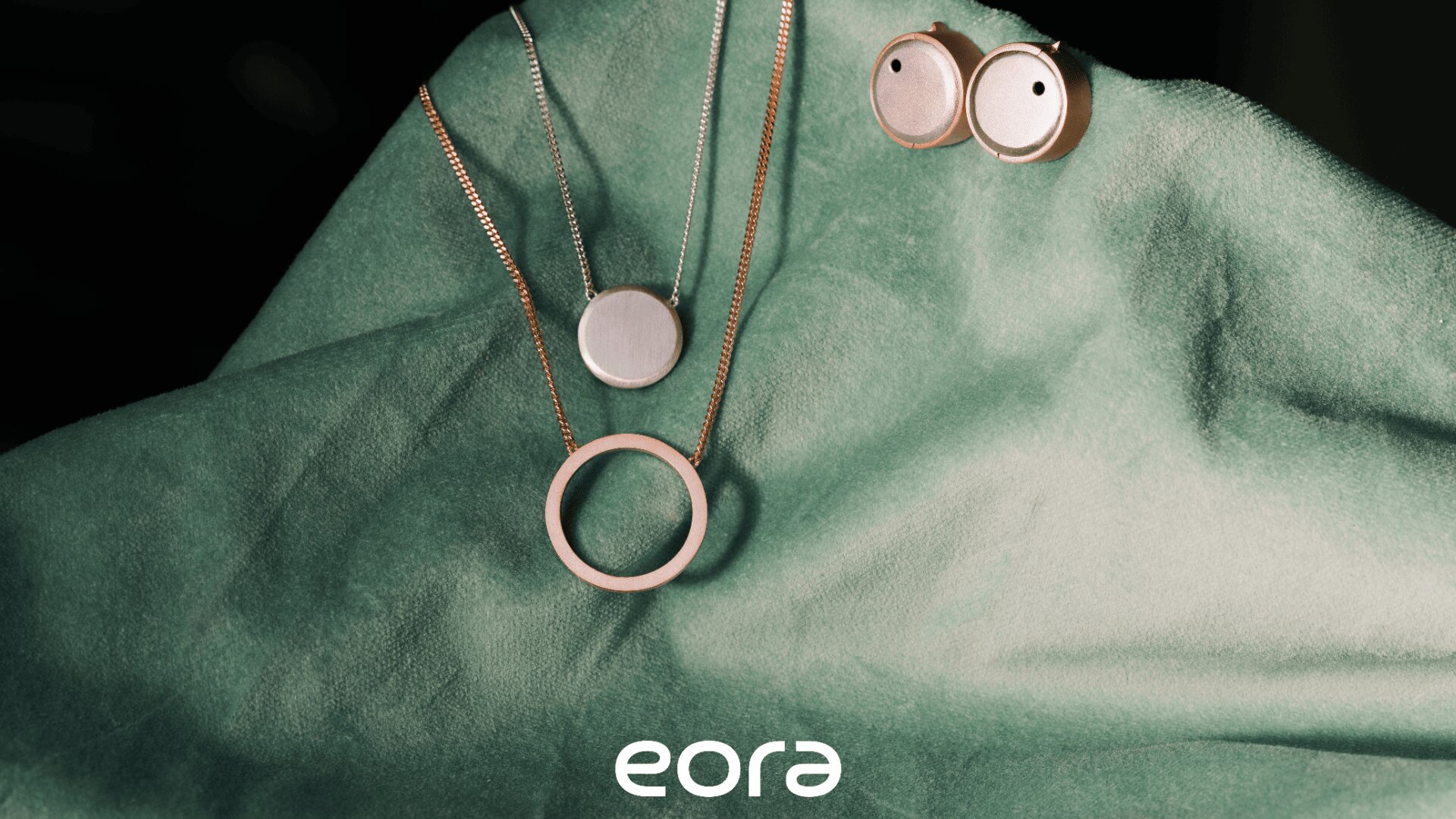 Minimalistischer Silberschmuck, darunter zwei Halsketten mit runden Anhängern und ein Paar runde Ohrstecker auf einem weichen grünen Stoff, mit dem Markennamen eora an der Unterseite.