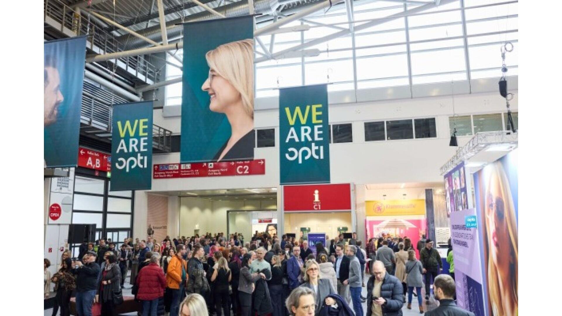 A large crowd gathers at an indoor expo or convention. Banners with a smiling woman’s profile and the text “WE ARE opti” hang from the ceiling. Bright lights and booths are visible in the background.