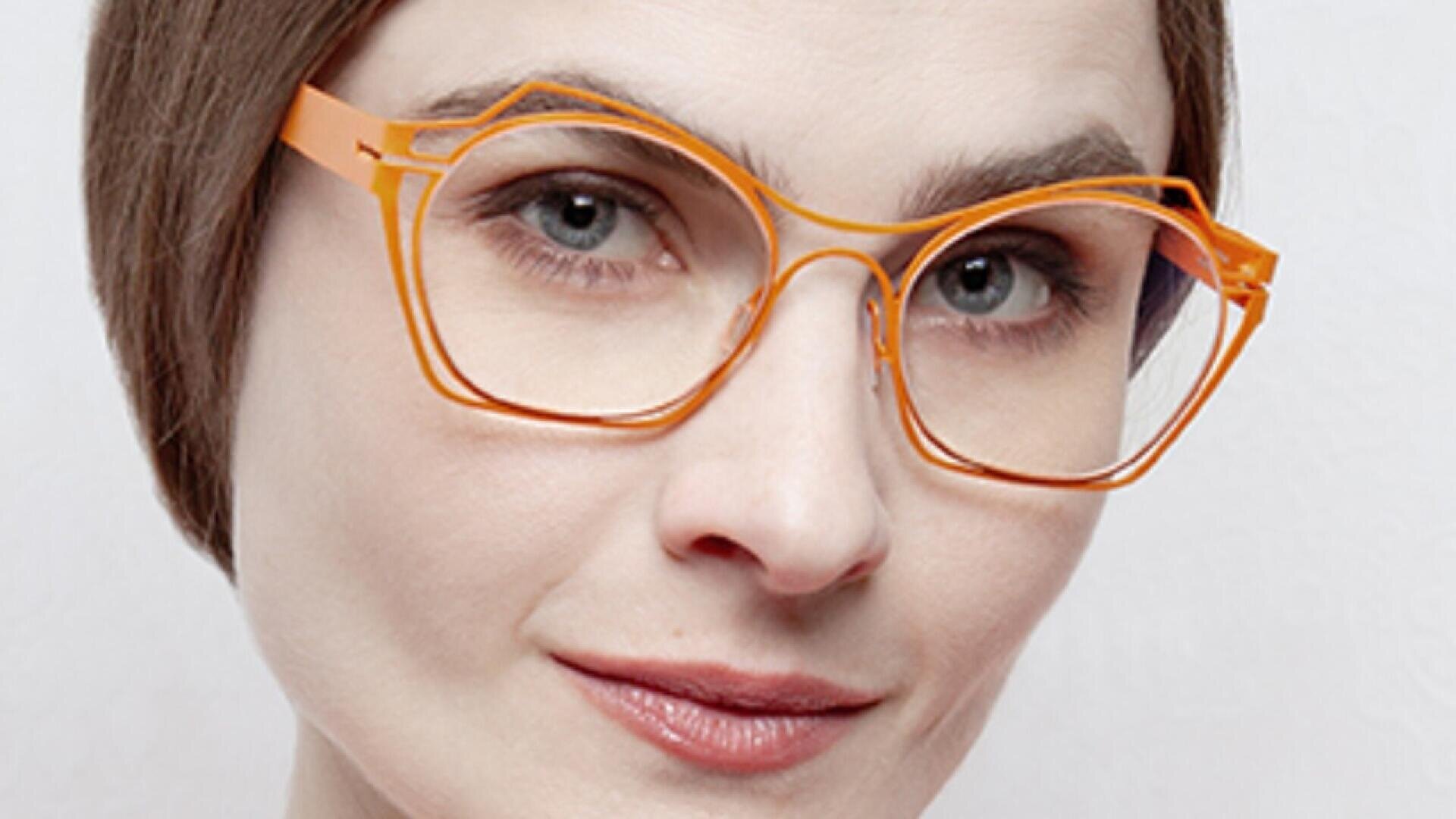 A person with light skin and short brown hair wears large, orange geometric eyeglasses and smiles slightly, looking directly at the camera.