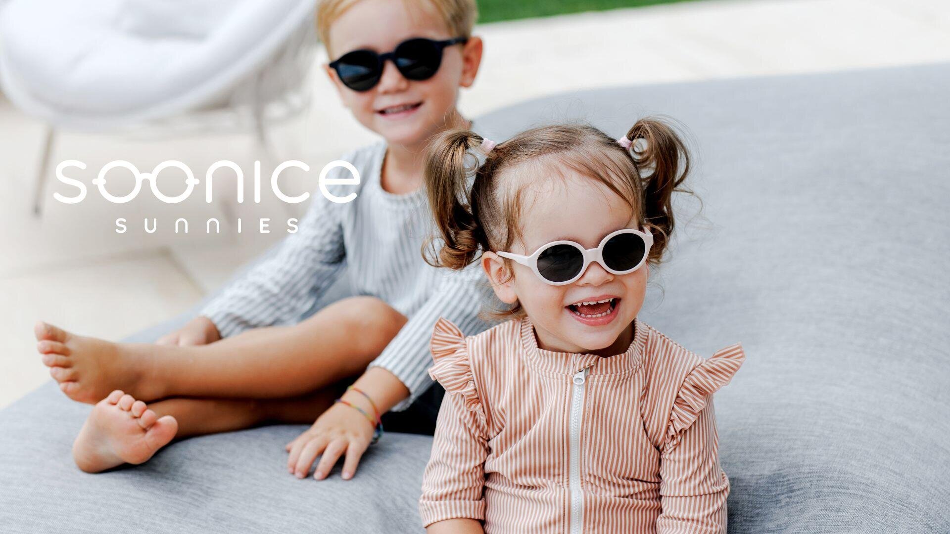 Zwei lächelnde kleine Kinder mit Sonnenbrille sitzen auf einem grauen Outdoor-Kissen. Das vordere Kind hat Zöpfe und trägt ein gestreiftes Outfit; das hintere Kind hat kurze Haare und trägt ein helles Outfit. Das Logo von SOONICE SUNNIES ist zu sehen.