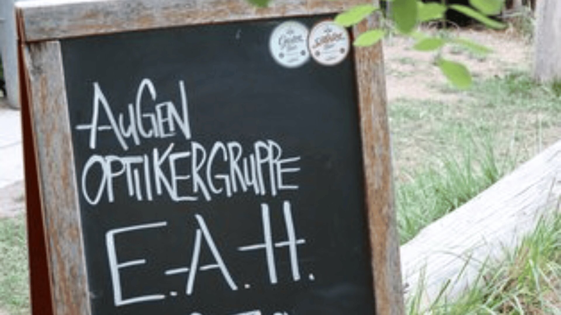 Ein rustikales Kreideschild im Freien zeigt handgeschriebenen Text in Deutsch: Augen Optikergruppe E.A.H. Im Hintergrund sind grünes Gras und Pflanzen zu sehen.