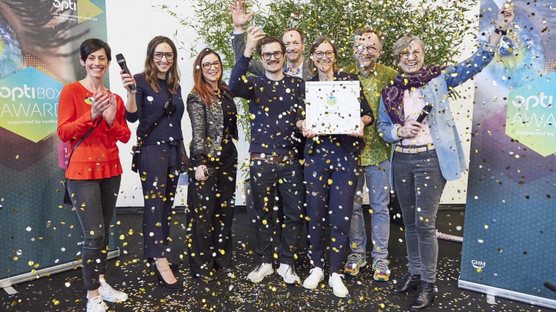 A group of nine people celebrate on stage with confetti falling around them; several are smiling, holding a trophy or certificate, and raising their arms in excitement at an award event.