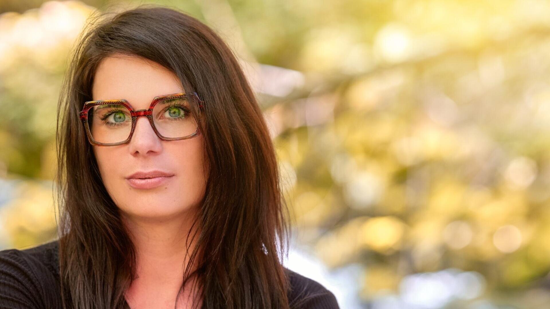 A woman with long dark hair and large, square glasses looks at the camera while standing outdoors. The background is softly blurred with warm, golden light and greenery.