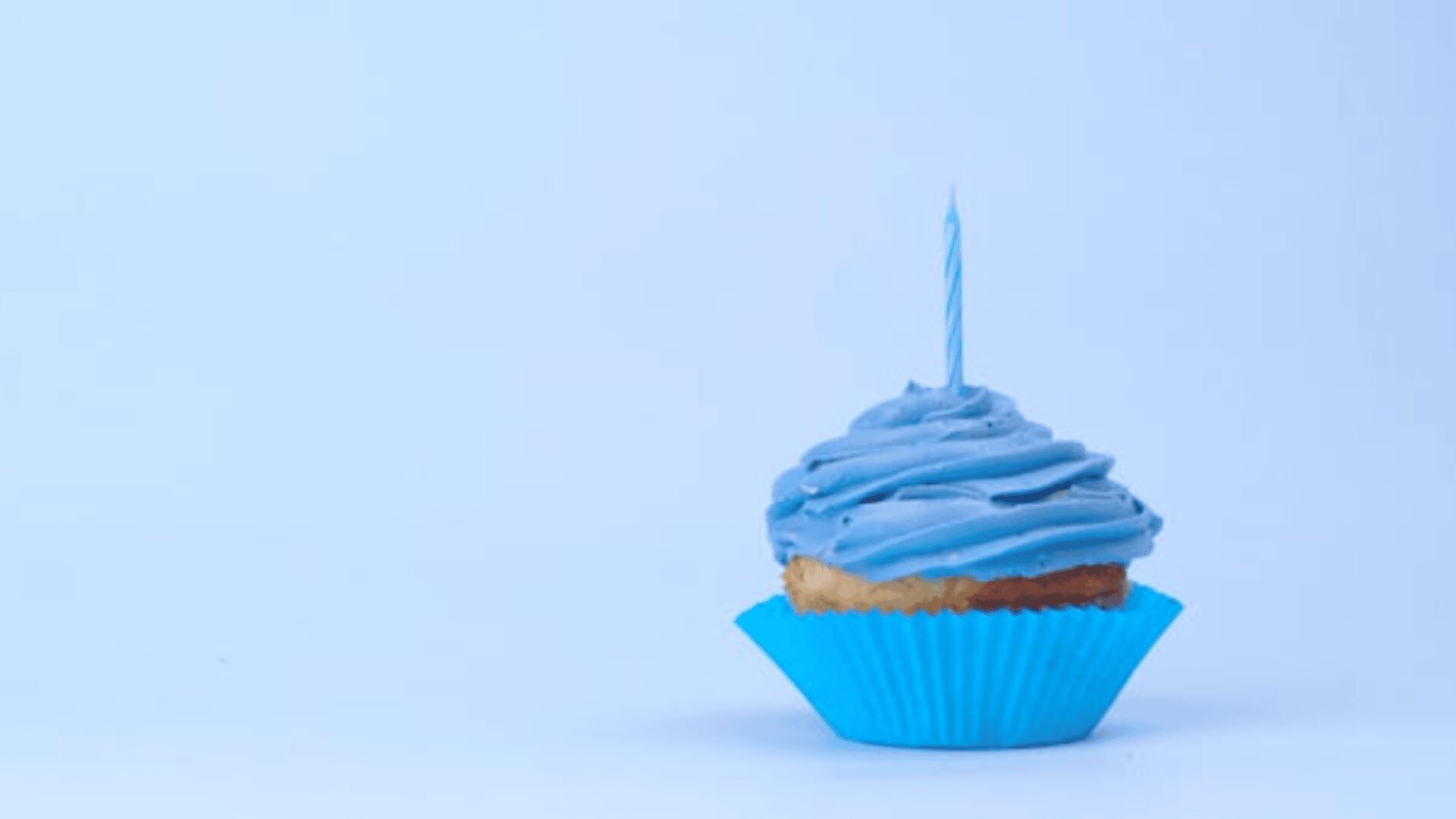 Ein einzelner Cupcake mit blauem Zuckerguss und einer brennenden blauen Geburtstagskerze, platziert in einem blauen Cupcake-Förmchen, wird vor einem schlichten hellblauen Hintergrund präsentiert.