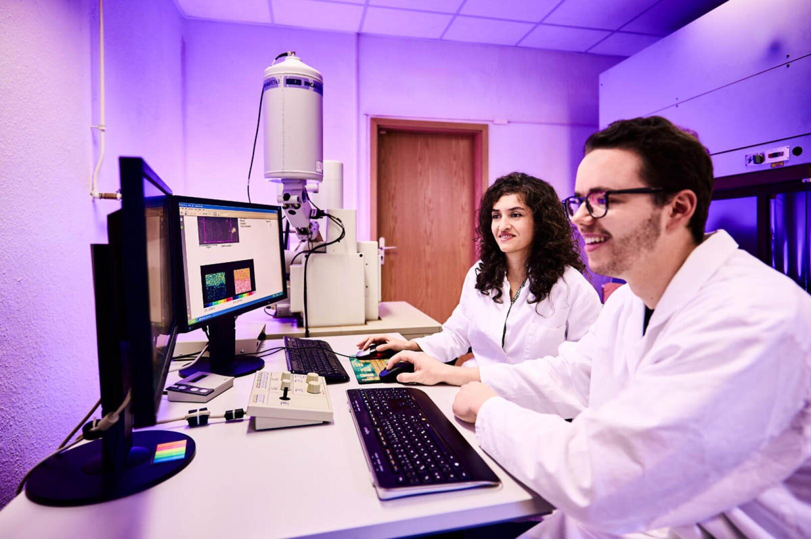 Two scientists in white lab coats sit at a desk in a laboratory, analyzing colorful images on computer monitors and using scientific equipment against a purple backdrop.