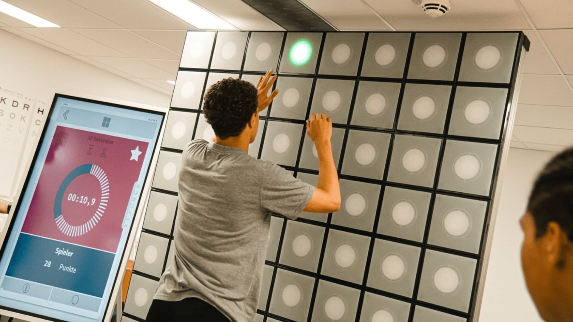 Eine Person berührt eine große Wand, die mit quadratischen Leuchttafeln bedeckt ist, wobei eine Tafel grün leuchtet. Ein digitaler Bildschirm in der Nähe zeigt einen Timer und Spielstatistiken an. Eine weitere Person beobachtet das Geschehen im Vordergrund.